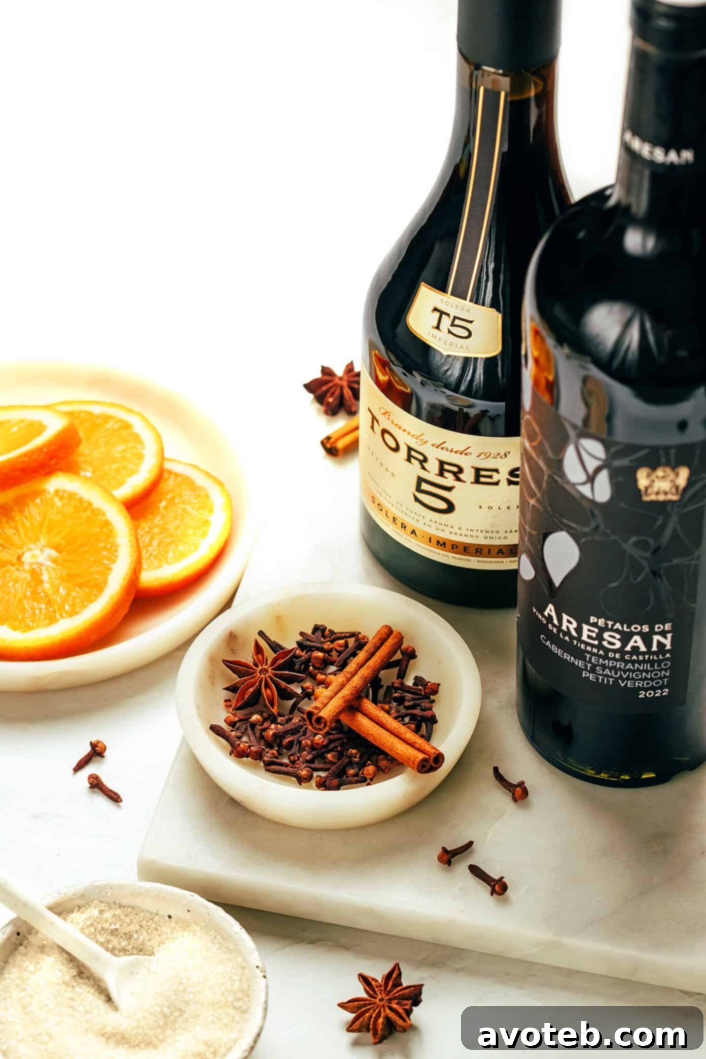A collection of essential mulled wine ingredients including a bottle of red wine, oranges, cinnamon sticks, star anise, and cloves, laid out on a wooden surface.