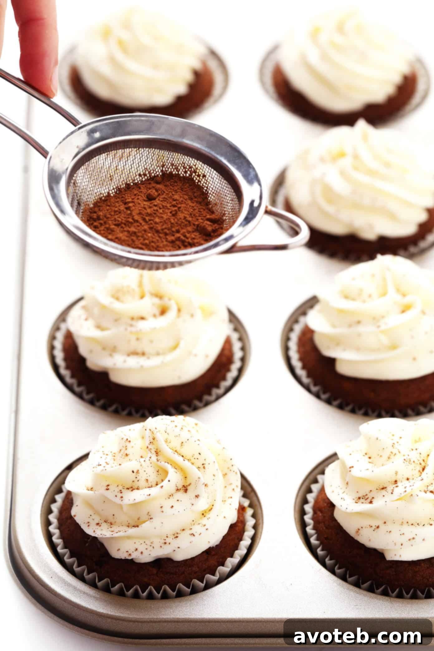 Hand sprinkling ground cinnamon over freshly frosted pumpkin cupcakes, enhancing their aromatic appeal.