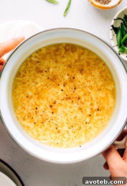 A beautifully presented bowl of Egg Drop Soup, garnished with green onions and a subtle swirl of sesame oil, on a white background.