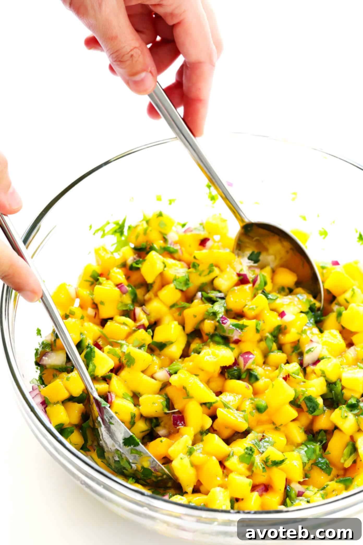 The BEST fresh mango salsa recipe! Made with just 5-ingredients, it's perfect as a dip or topping for your favorite Mexican dishes. Hands gently tossing diced mangoes, red onion, jalapeño, cilantro, and lime juice in a clear glass mixing bowl, illustrating the salsa-making process
