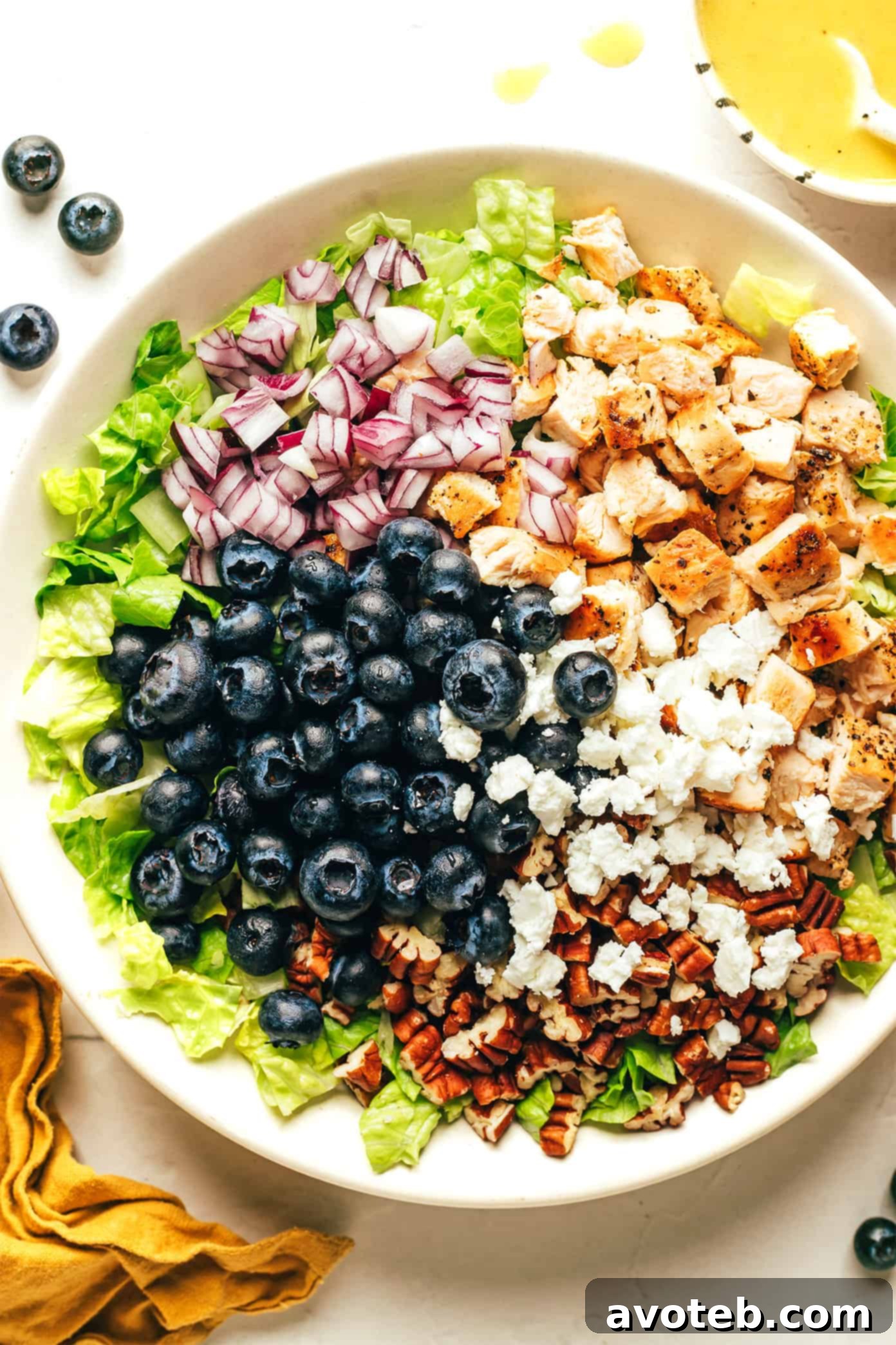 A large bowl filled with ingredients for Blueberry Chicken Chopped Salad, including chopped lettuce, red onion, pecans, and feta cheese, with fresh blueberries ready to be added.