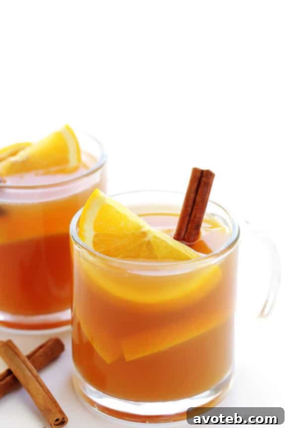 Two mugs of warm slow cooker apple cider, beautifully garnished with cinnamon sticks and orange slices, placed on a rustic table amidst fall decorations, ready for enjoyment.