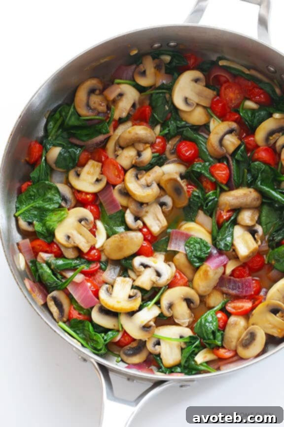 Overhead shot of a large skillet filled with sautéed mushrooms, red onions, garlic, cherry tomatoes, and wilting spinach, ready to be tossed with pasta.