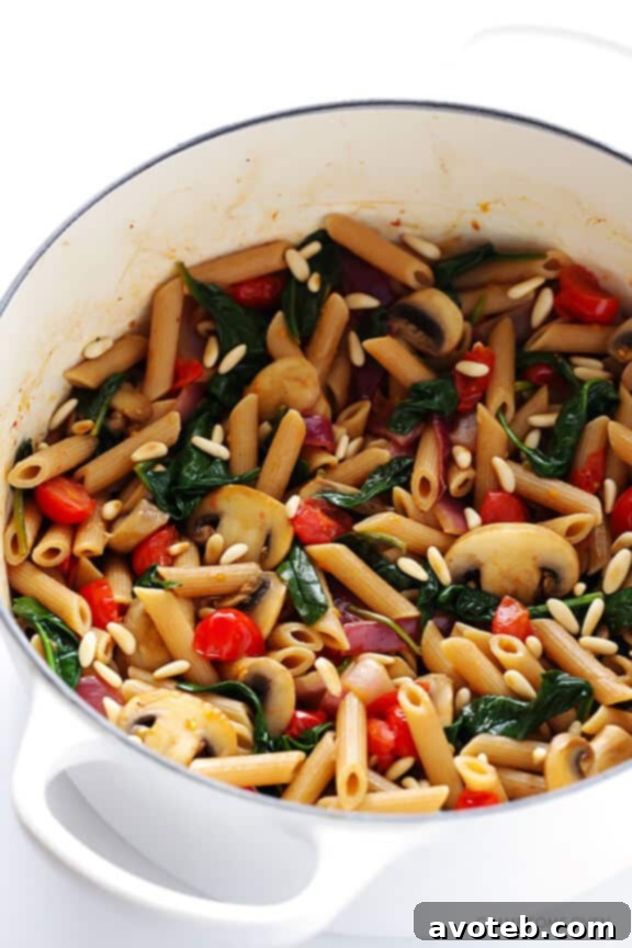 A serving of freshly cooked Whole Wheat Pasta with Mushrooms, Tomatoes, and Spinach, topped with toasted pine nuts and a sprinkle of Parmesan cheese.