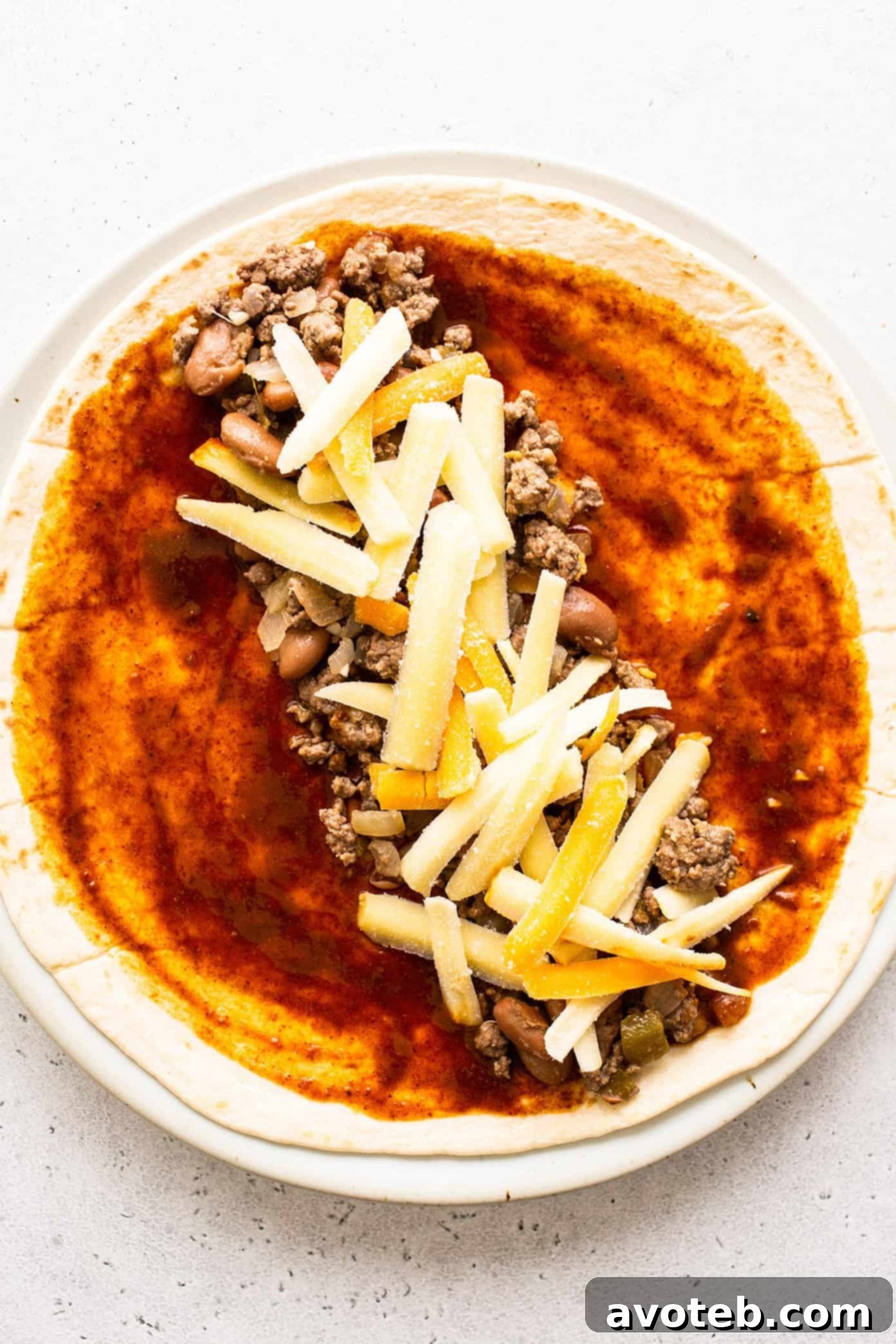 Beef enchilada filling spread on a tortilla with cheese and red enchilada sauce