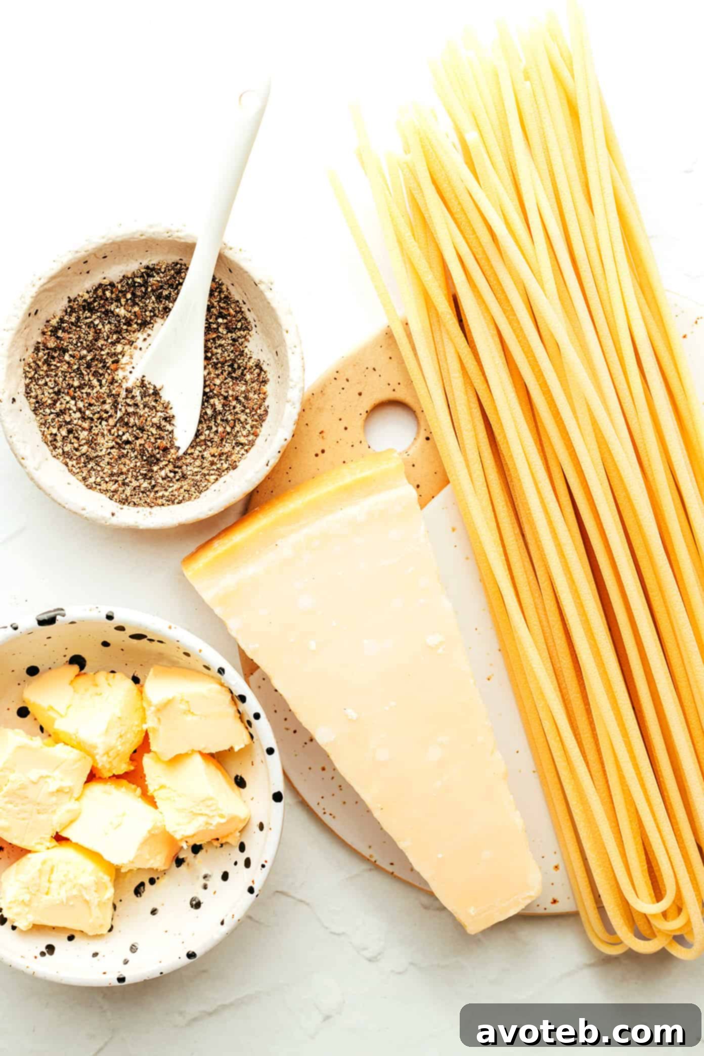 Cacio e Pepe ingredients laid out on a wooden board: bucatini pasta, block of Pecorino Romano cheese, whole black peppercorns, and butter cubes.