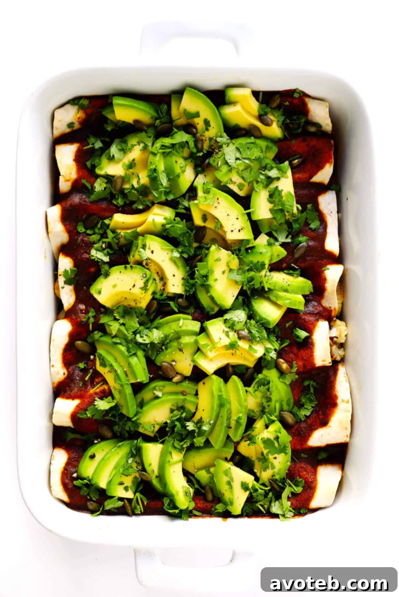 A close-up of a freshly baked pan of Roasted Cauliflower Enchiladas, generously adorned with vibrant green avocado slices and cilantro.