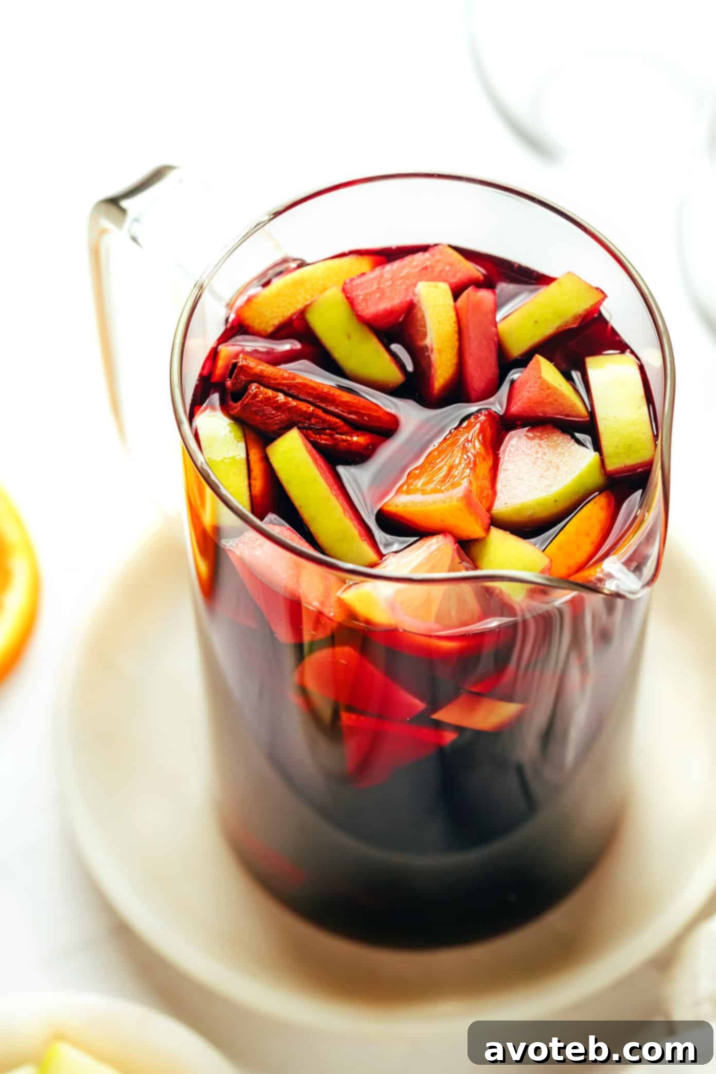 Close-up of a hand reaching for a glass of red sangria, with a pitcher full of fruit and ice in the background.