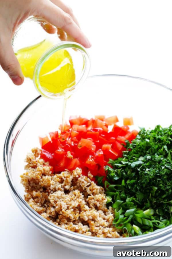 Hands tossing finely chopped fresh parsley, mint, tomatoes, scallions, and softened bulgur in a large bowl, preparing for the lemon vinaigrette.