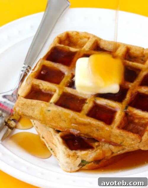 Close-up of a stack of Zucchini Waffles with a pat of melting butter and maple syrup, showing the fluffy texture and green zucchini flecks.