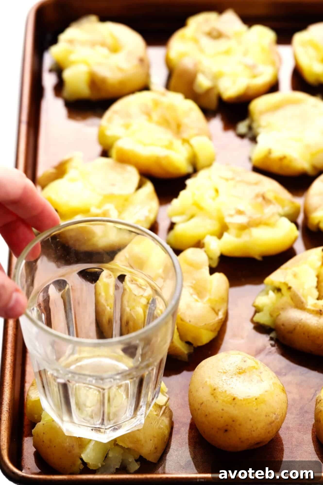 Crispy Smashed Potatoes 4 A close-up image of a hand using a drinking glass to gently smash boiled potatoes on a baking sheet.