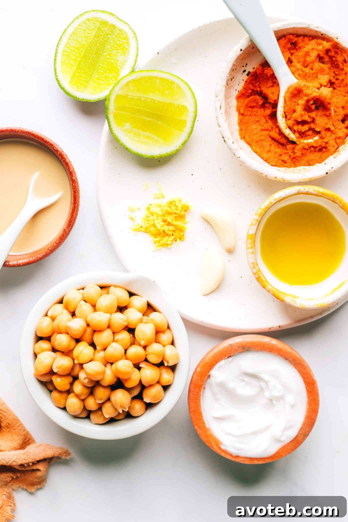 A selection of fresh ingredients including chickpeas, tahini, coconut milk, lime, ginger, garlic, and red curry paste laid out for making hummus.