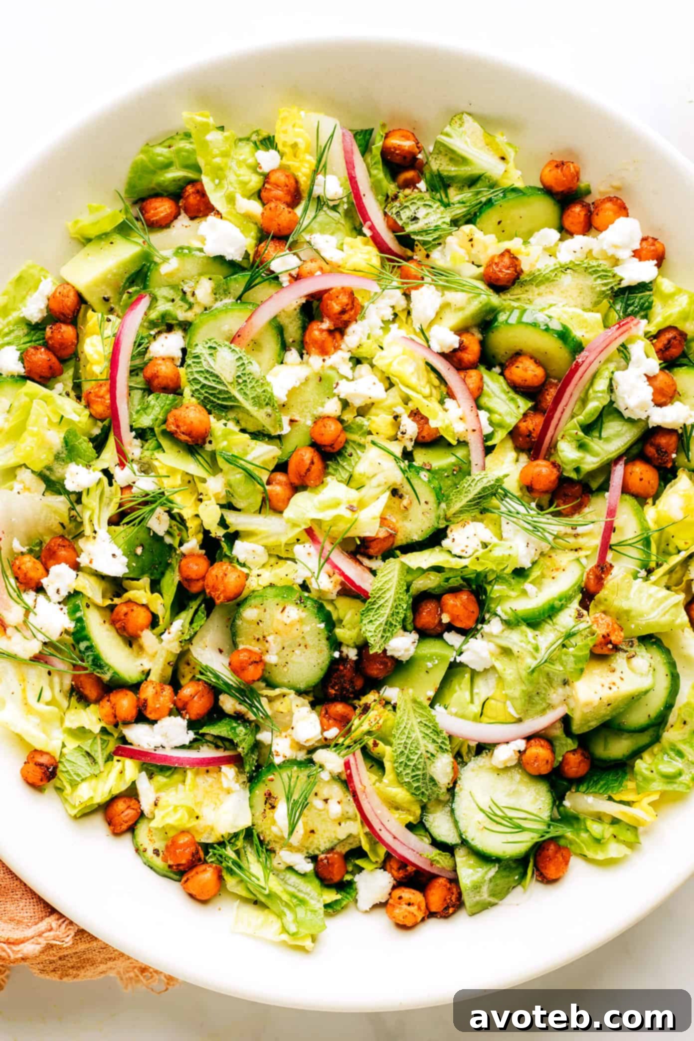A beautifully presented roasted chickpea, avocado, and cucumber salad, highlighting its fresh ingredients and vibrant colors. The salad is in a white bowl, garnished with fresh herbs, against a rustic background.