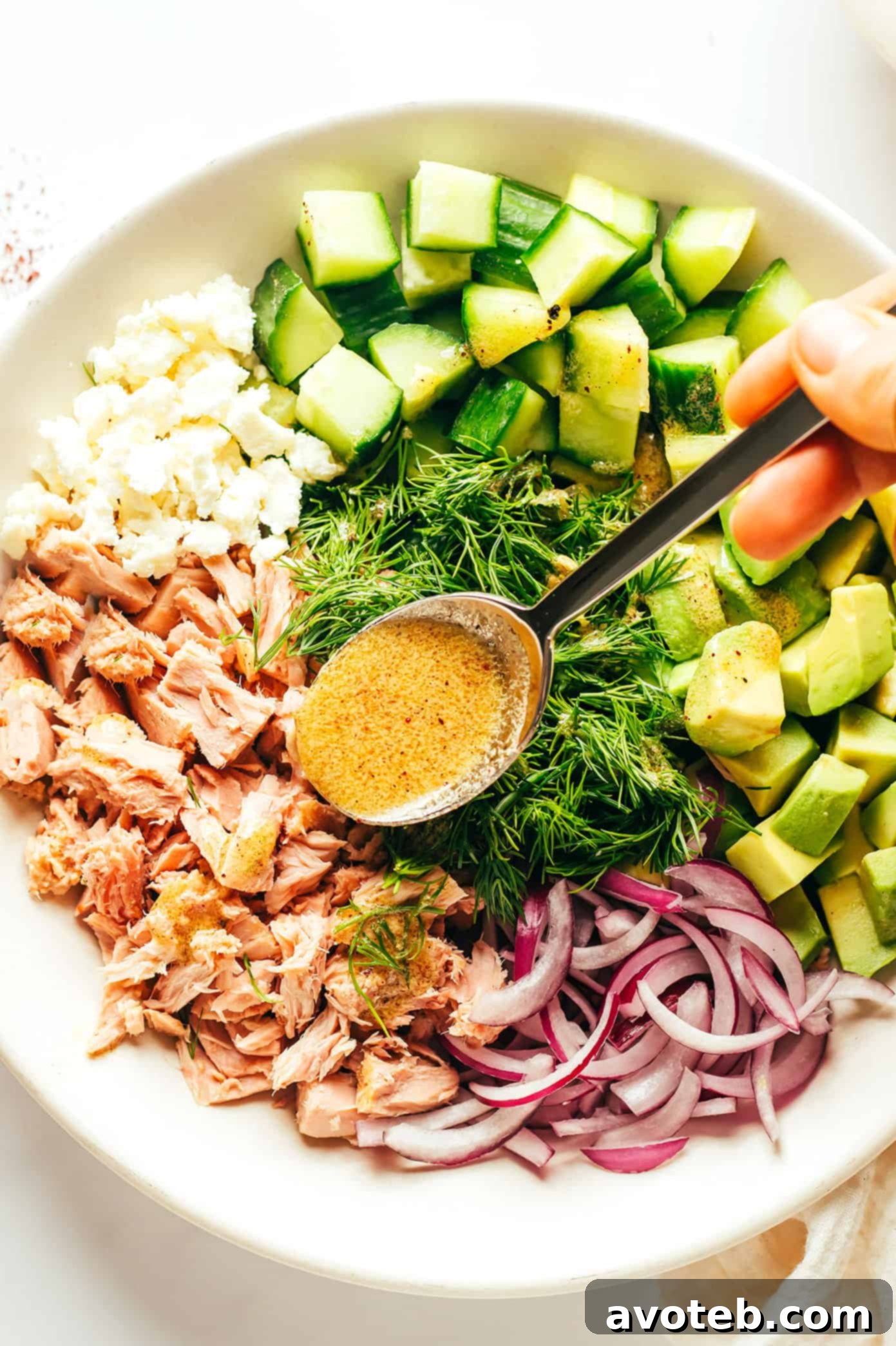 Adding vibrant lemon-sumac dressing to a bowl of fresh Tuna Avocado Salad ingredients before tossing.