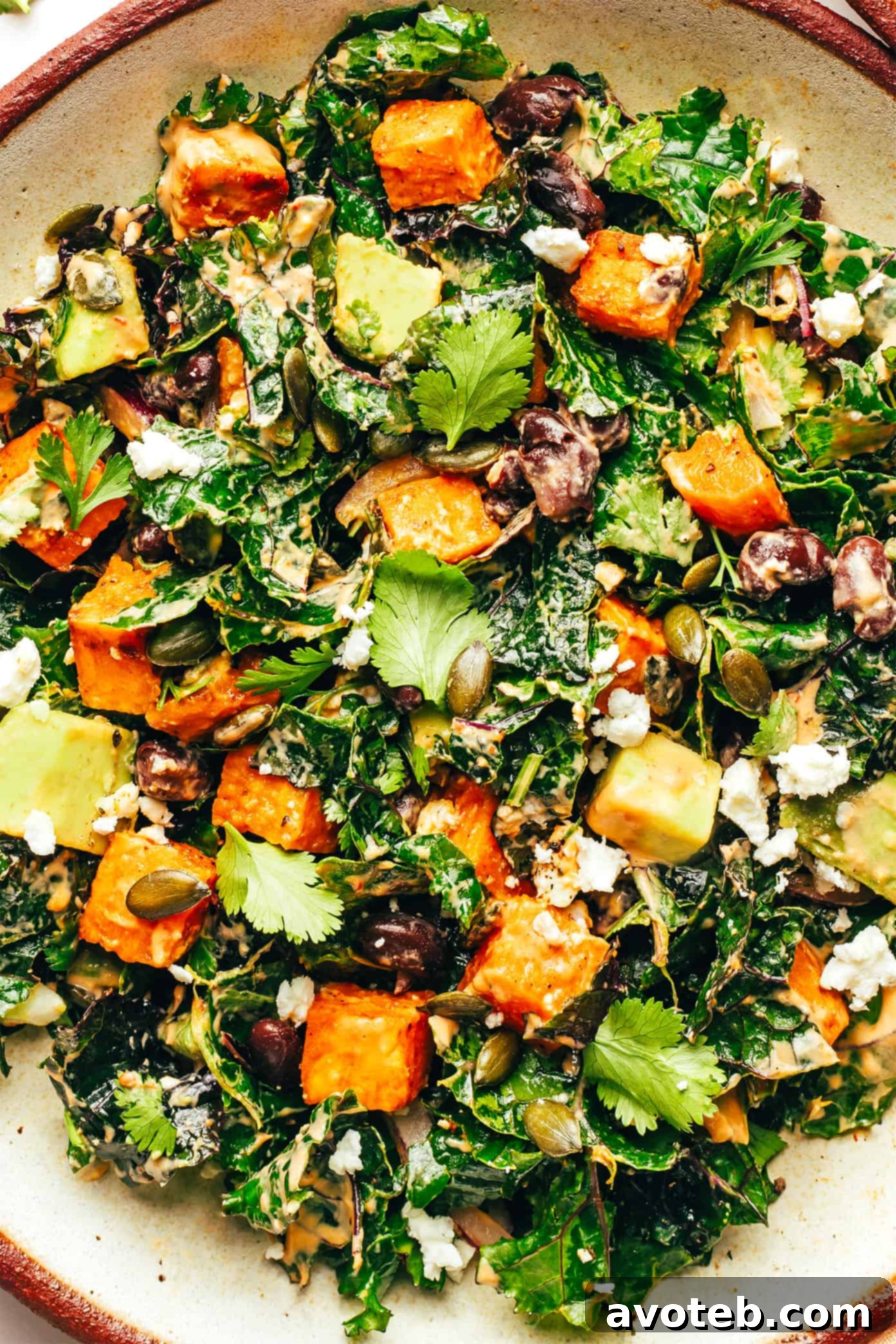 Close-up shot of a bowl of Roasted Sweet Potato Avocado Kale Salad, garnished with avocado slices, pepitas, and a drizzle of chipotle tahini dressing, ready to be eaten with a fork.