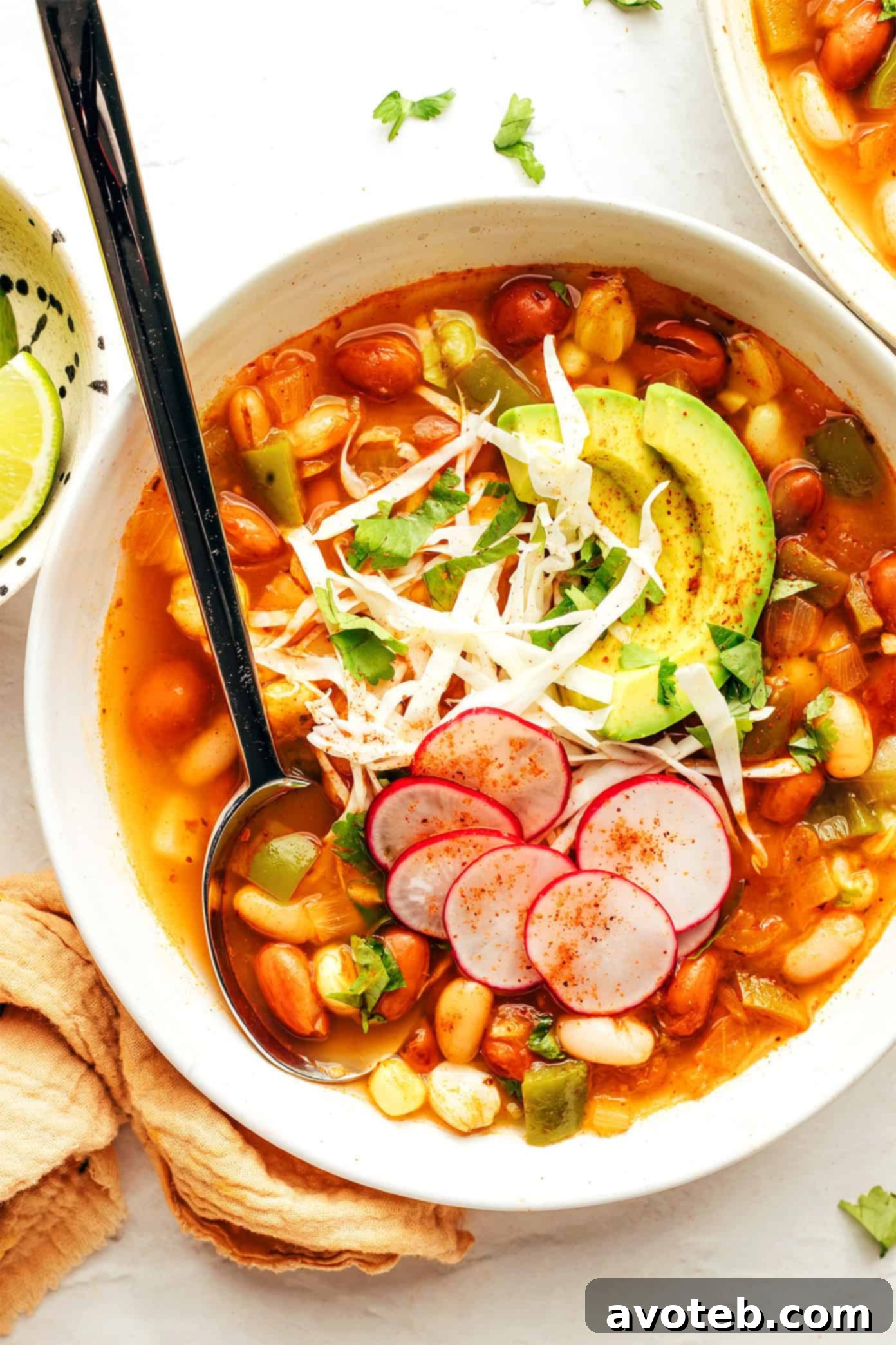 A side view of a bowl of vegetarian pozole, richly colored and garnished, with a spoon nestled in.