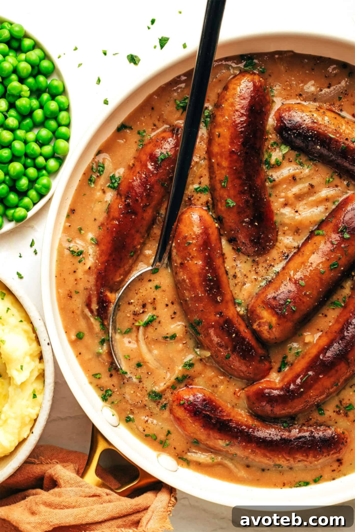 A generous serving of bangers and mash on a plate, featuring two sausages, a large scoop of mashed potatoes, and a pool of dark, glossy onion gravy, garnished with parsley.