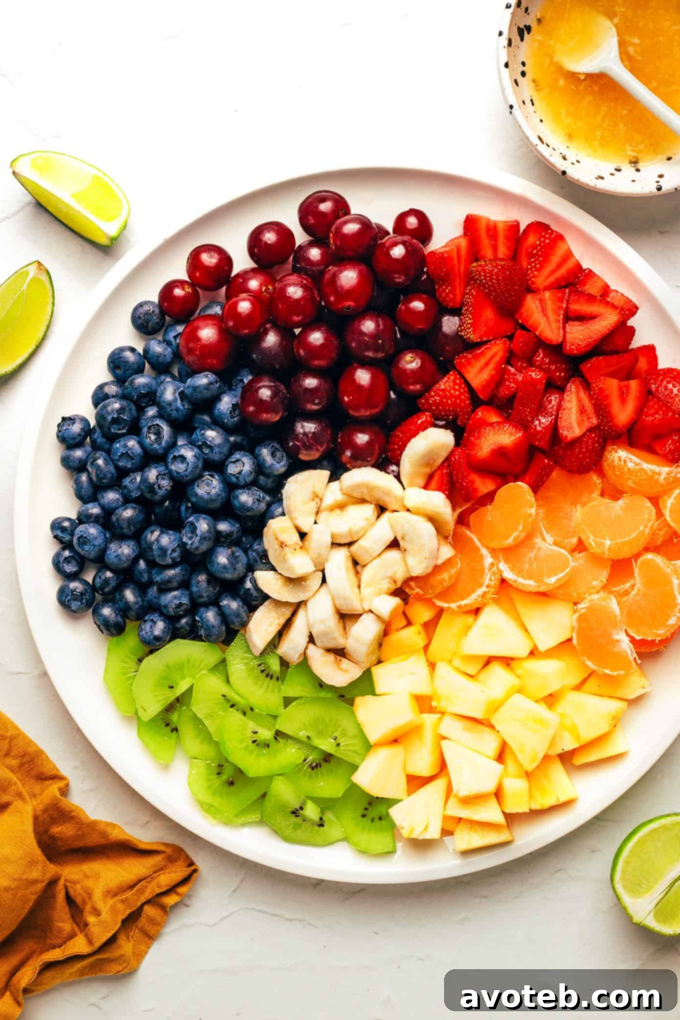 Ingredients for fruit salad laid out: fresh pineapple chunks, sliced strawberries, blueberries, red grapes, mandarin oranges, peeled kiwis, and bananas. Also, small bowls of lime juice, honey, and fresh ginger for the dressing.