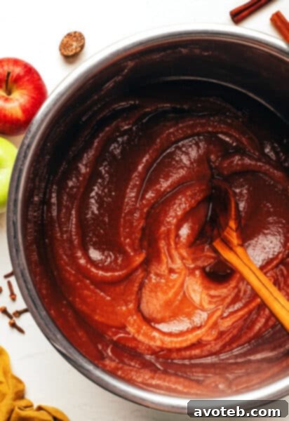 A jar of homemade apple butter, rich brown in color, with a spoon resting on the side, and a slice of bread spread with apple butter.